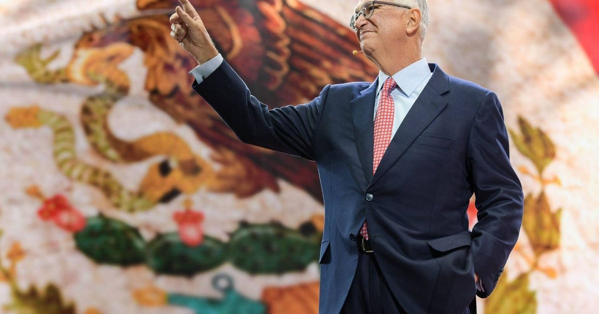 Persona en traje oscuro señalando, con emblema de México de fondo en evento público.