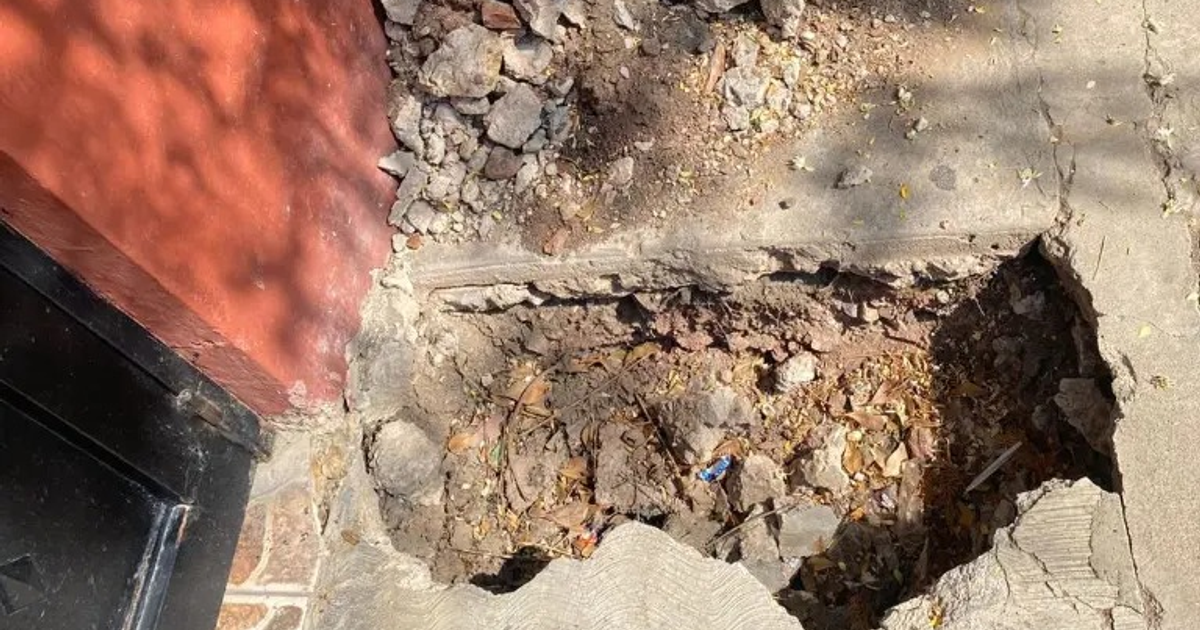 Agujero en banqueta con piedras sueltas y desechos, pared roja y puerta negra al fondo.