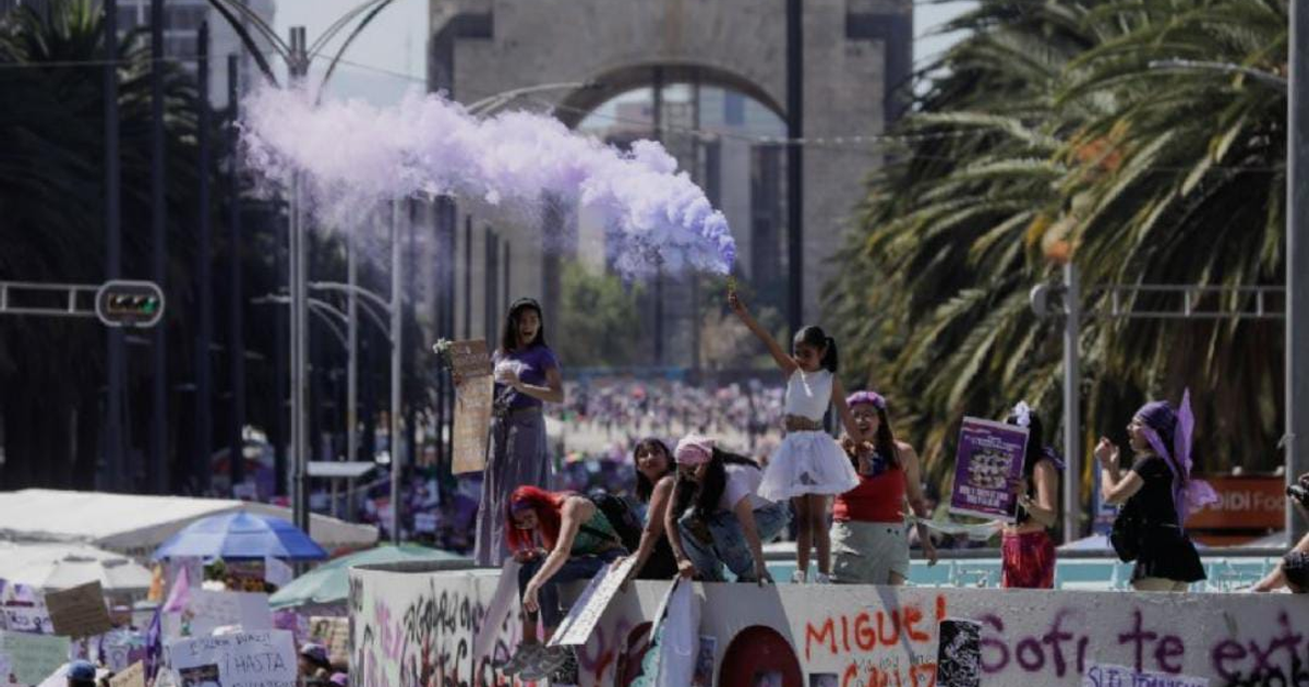 Mujeres sosteniendo pancartas en la marcha por el 25N en Ciudad de México entre humo morado y palmeras.