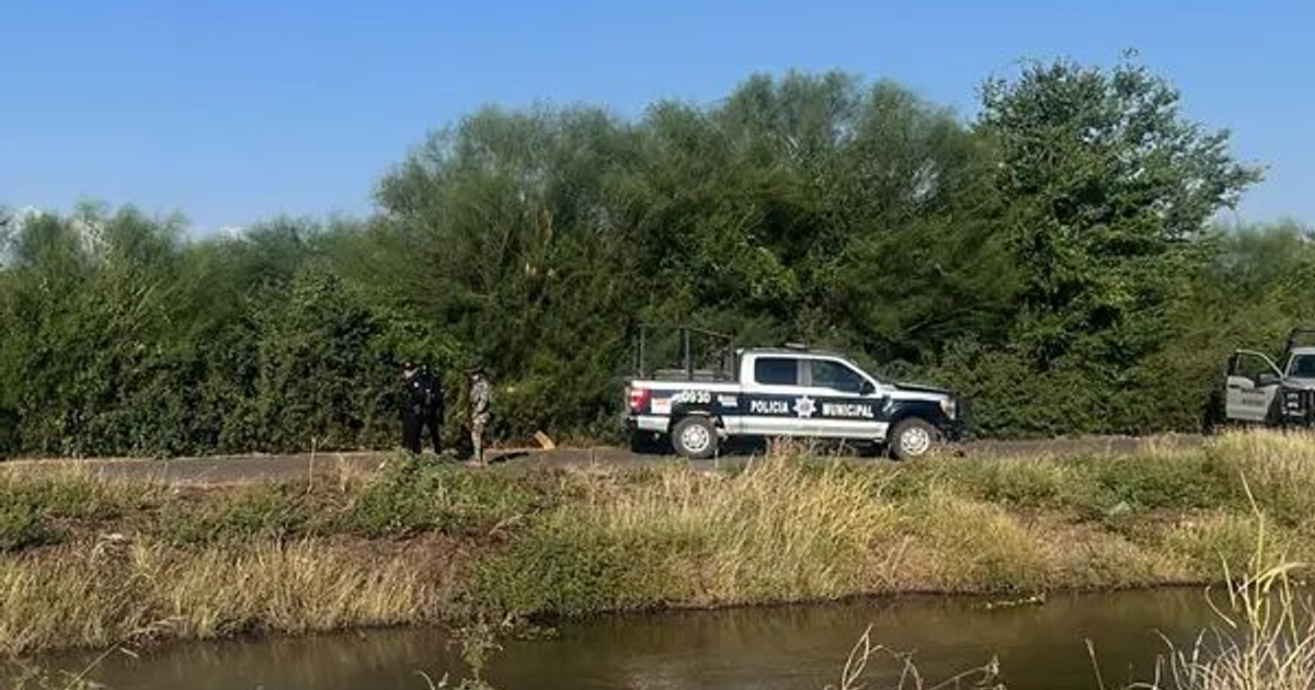 Vehículo de Policía Municipal al lado de un canal con dos personas de pie, búsqueda en curso de niño desaparecido, cielo despejado.