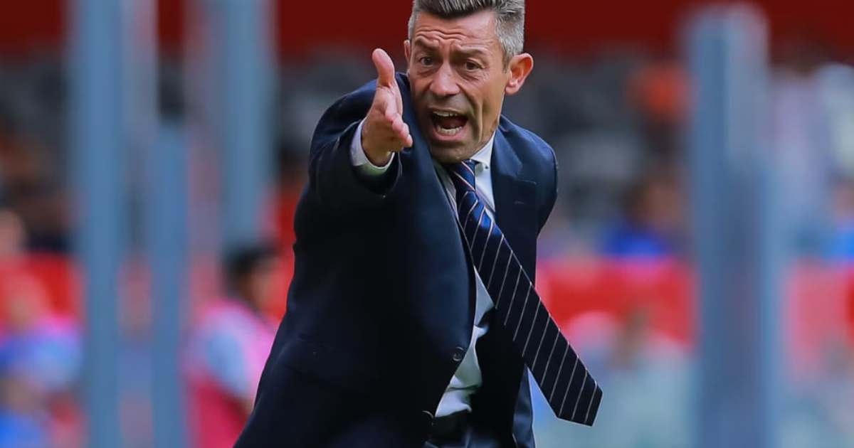 Pedro Caixinha dando instrucciones en un evento deportivo, vestido con traje oscuro y corbata, con espectadores al fondo.