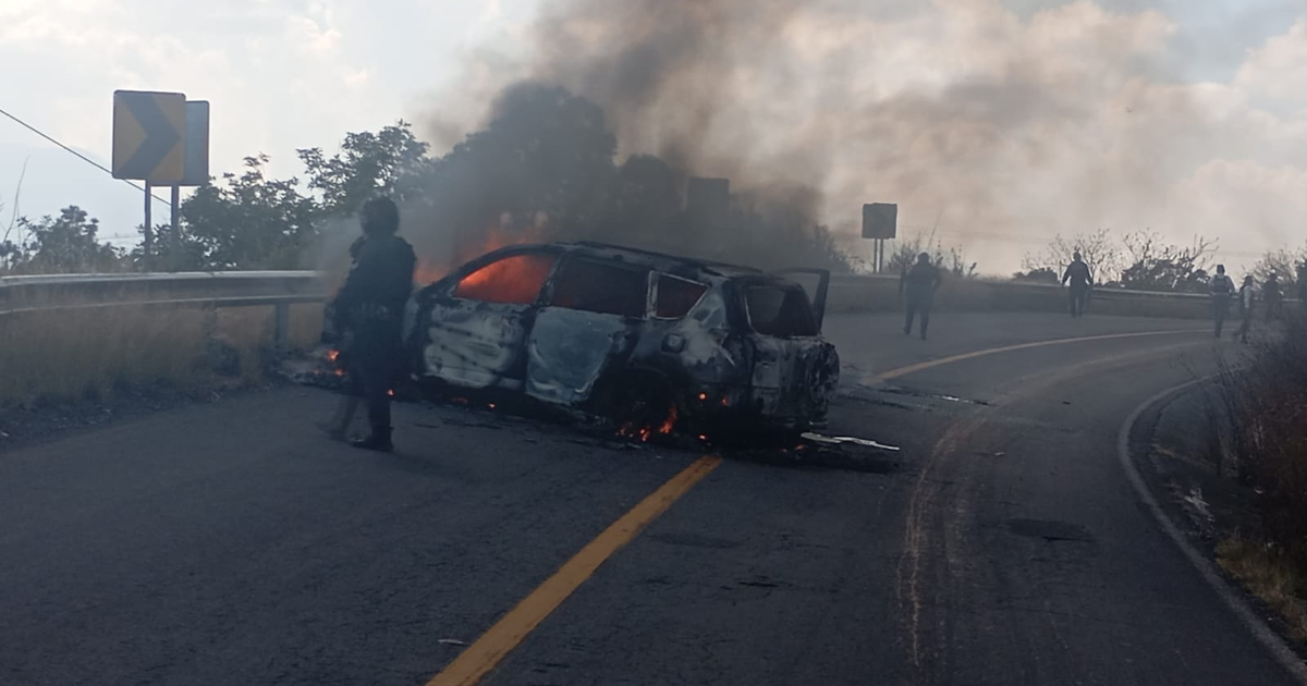Automóvil en llamas en carretera con humo y personas alrededor, reflejando narcobloqueos en Michoacán.