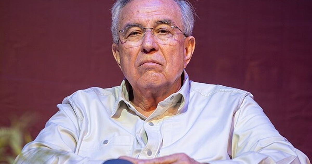 Hombre mayor con gafas y expresión seria, fondo burdeos