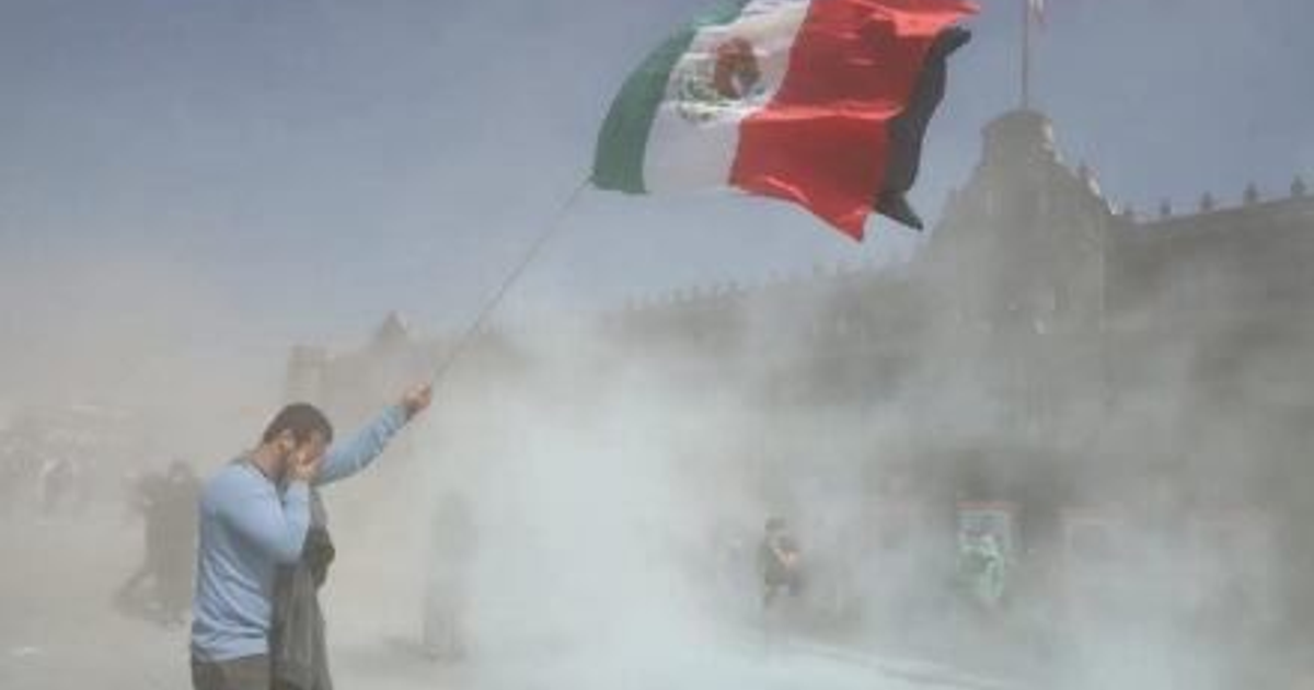 Persona con bandera de México en marcha, rodeada de polvo, frente a un edificio.