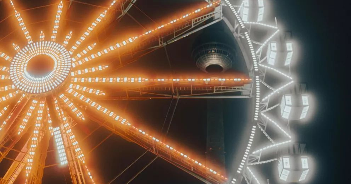 Noria iluminada en el Festival de la Cosecha, New Roads, con luces brillantes y una estructura alta al fondo.