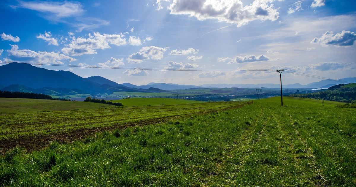 Vista de un paisaje rural con hierba verde, montañas al fondo, cielo azul con nubes y líneas eléctricas cruzando el escenario.