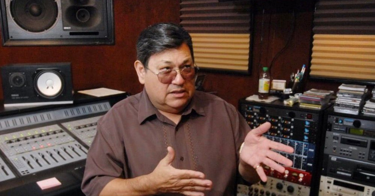 Persona en estudio de grabación simbolizando a Abraham Quintanilla, rodeado de equipo de audio, CDs y accesorios de oficina.