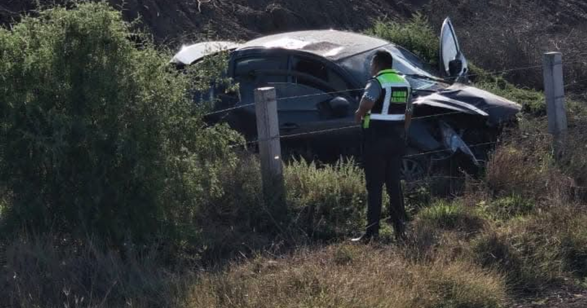 Auto negro accidentado junto a carretera y policía investigando, rodeado de vegetación.