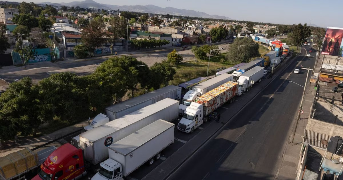 Camiones de carga estacionados en carretera urbana con edificios, montañas y tráfico denso al fondo.