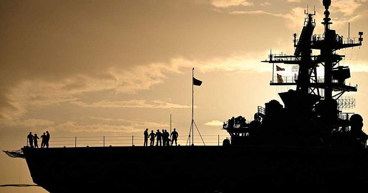 Silueta de portaaviones al atardecer con personal en la cubierta y bandera ondeando, fondo dorado