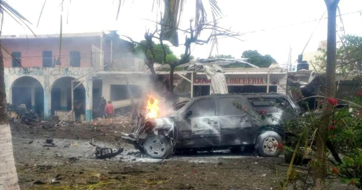Vehículo destruido en la explosión de Coahuayana, Michoacán, emitiendo llamas y humo con daños en edificios de fondo.
