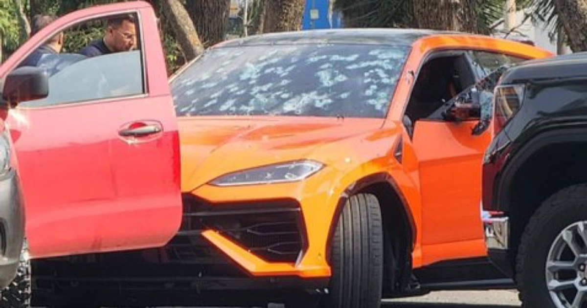 Lamborghini Urus naranja con ventana rota junto a camioneta roja, rodeado de personas en una escena de bala.