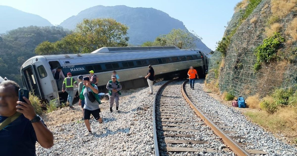 Escena de un tren descarrilado en un paisaje montañoso natural, con múltiples personas alrededor.