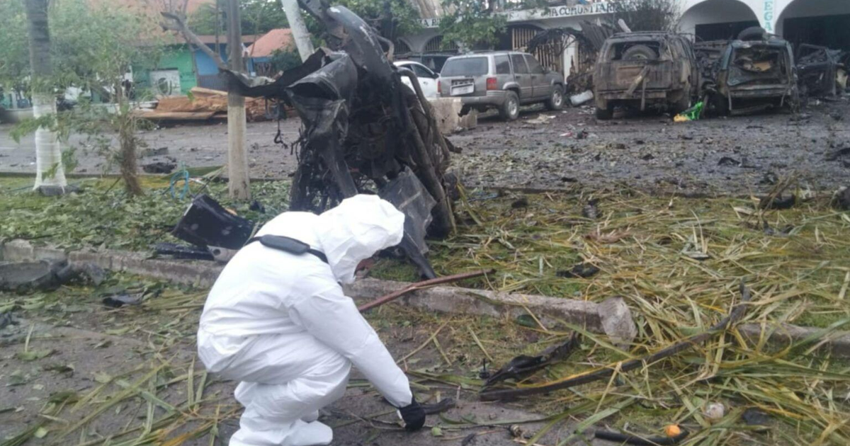 Investigador en traje protector examina la escena de explosión en Coahuayana, Michoacán, entre vehículos dañados y escombros.