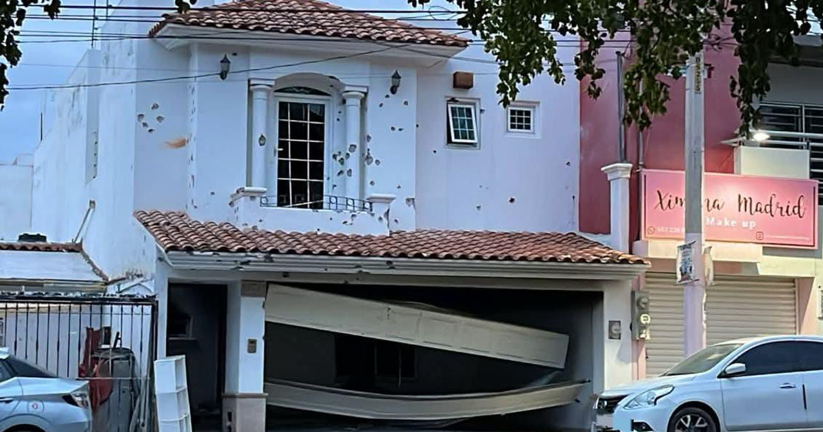 Casa con daños en Culiacán tras ataque armado: fachada blanca con impactos y garaje afectado. Negocio adyacente y automóviles en la calle.