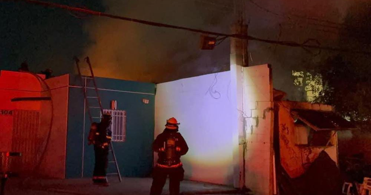 Dos bomberos combatiendo un incendio con humo espeso en Culiacán, junto a una escalera apoyada en una pared azul en un ambiente con luces rojas.