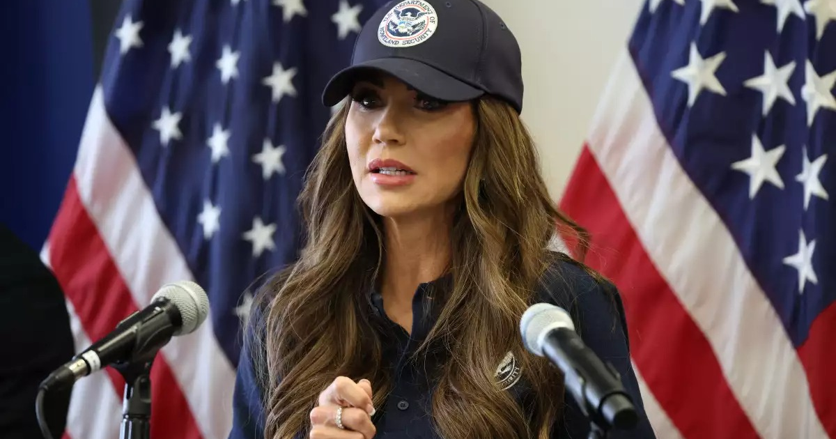 Kristi Noem habla ante micrófonos con banderas estadounidenses tras ella.