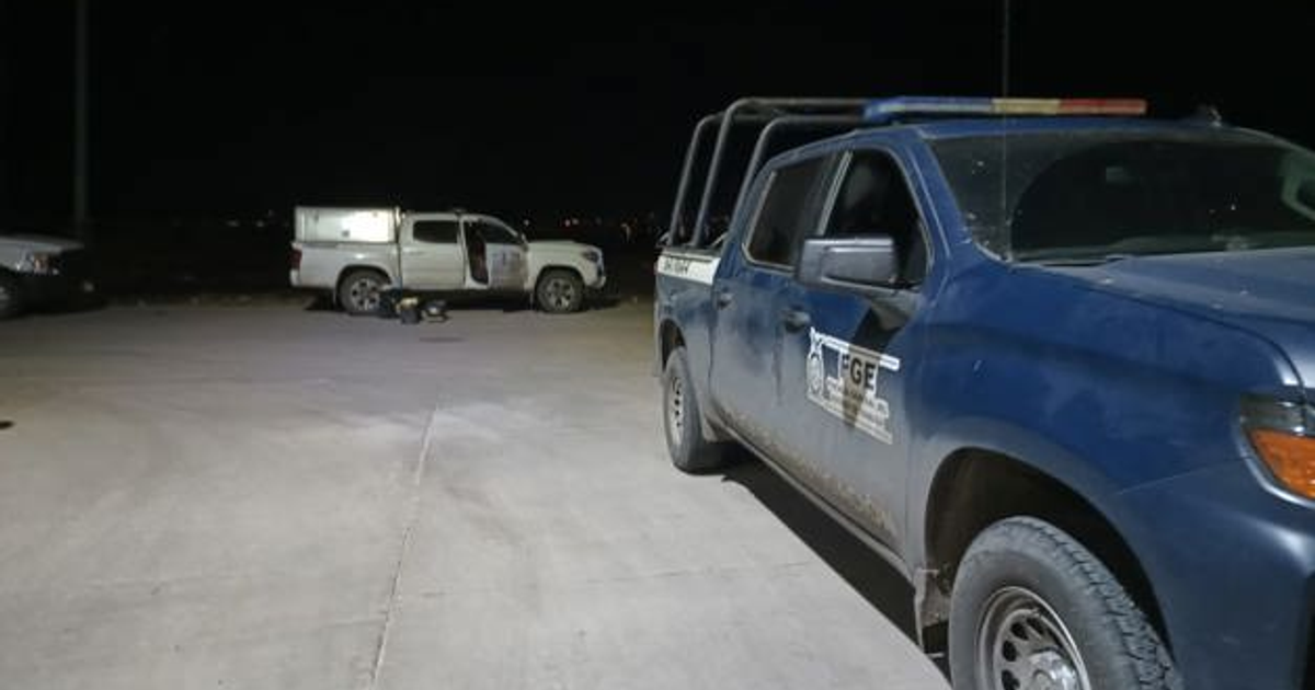 Dos vehículos estacionados en un área oscura, uno azul con insignias y otro blanco con la puerta abierta, en un lugar desierto, posiblemente conectado con el incidente en Culiacán.