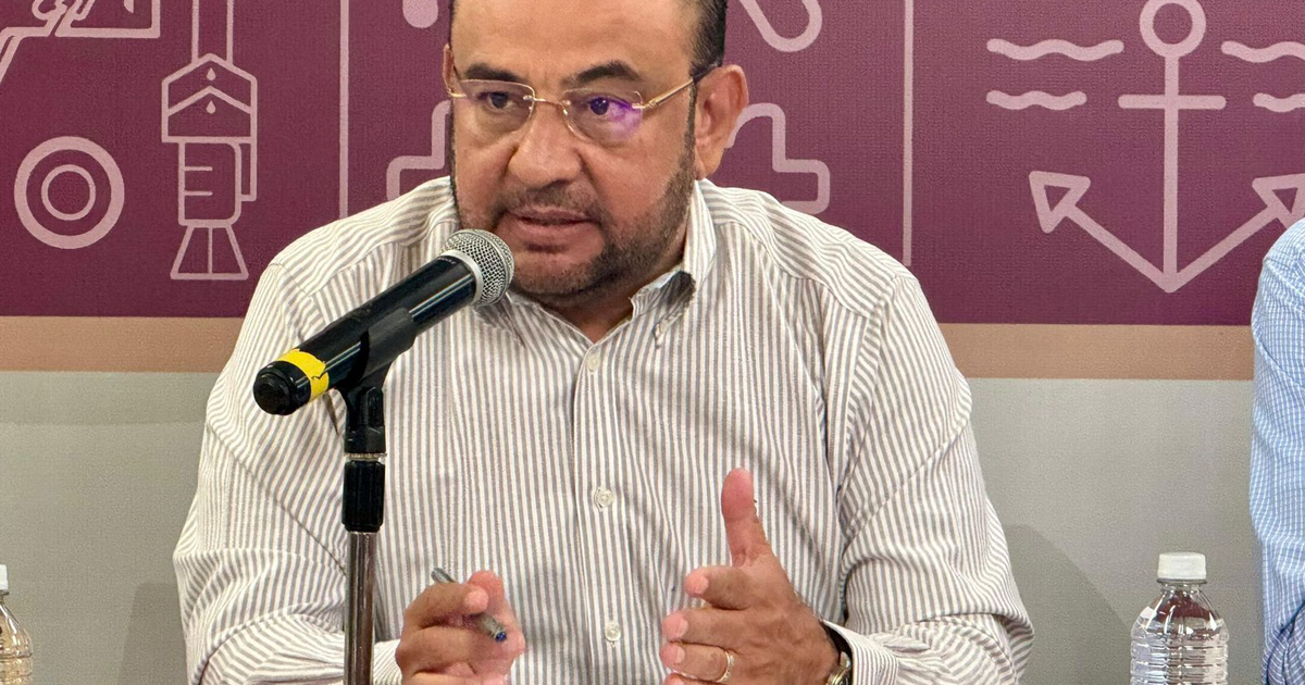 Marcos Osuna hablando en evento con micrófono y camisa de rayas, tema marino de fondo.