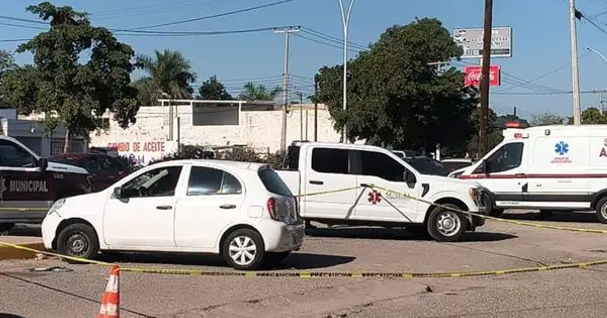 Escena de emergencia acordonada frente a un hospital con vehículos policiales y de emergencia, árboles y un edificio al fondo.