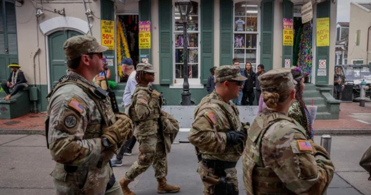 Fuerzas armadas en uniforme militar patrullando en Nueva Orleans con tiendas de fondo y público observando.