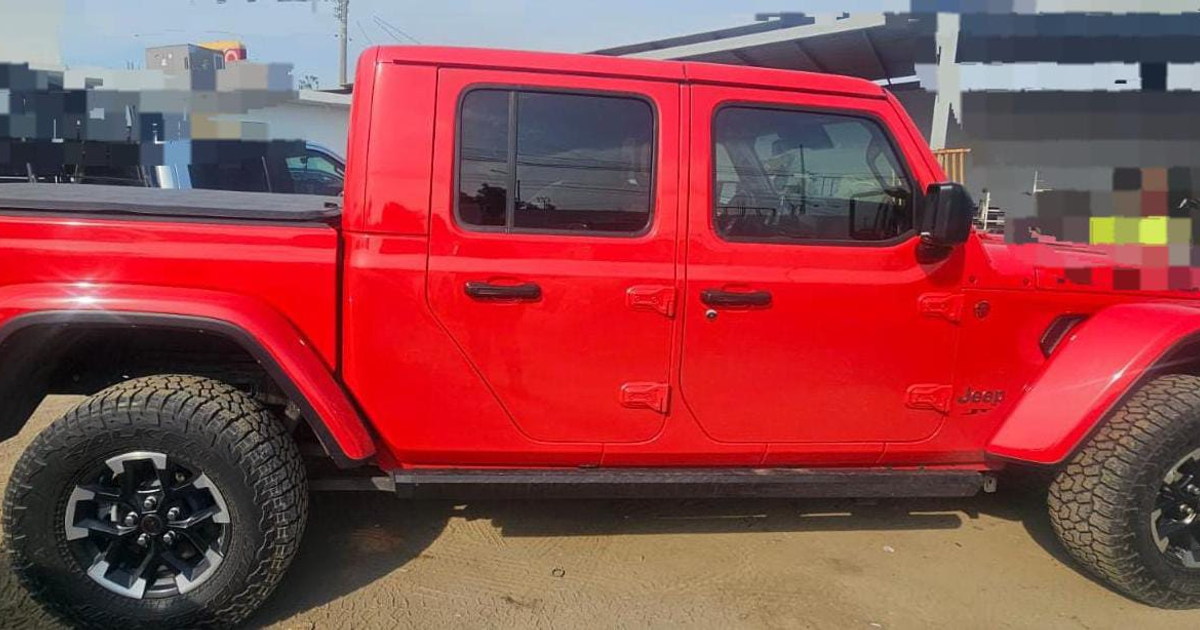 Camioneta Jeep roja con estructura de varios asientos, en un entorno de tierra, acompañada por otros vehículos y estructura al fondo.