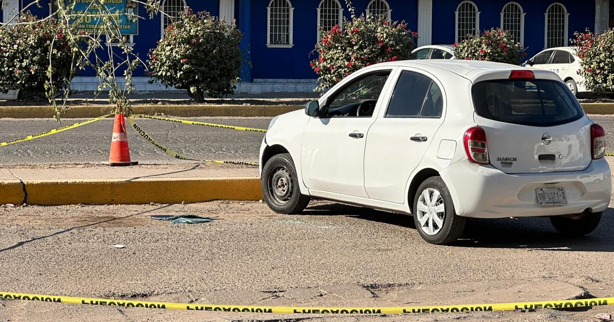 Nissan March blanco estacionado con cinta amarilla de seguridad y neumático dañado, escena de investigación detrás de un edificio azul.