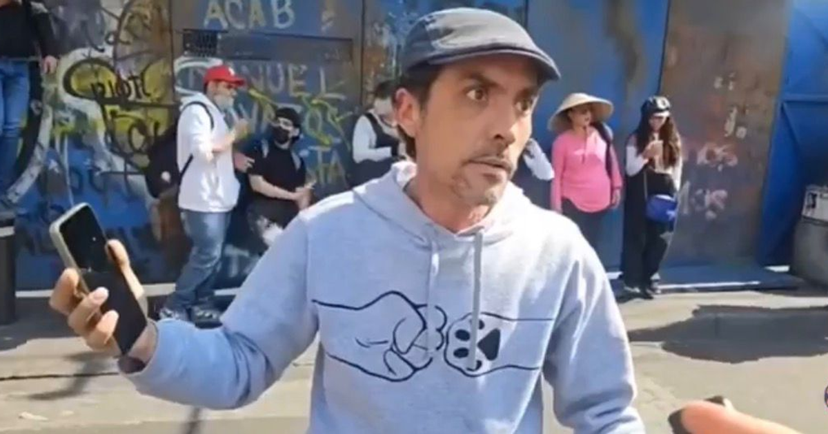 Hombre joven con teléfono móvil delante de un mural de graffitis, ciudadanos al fondo, algunos con mascarillas.