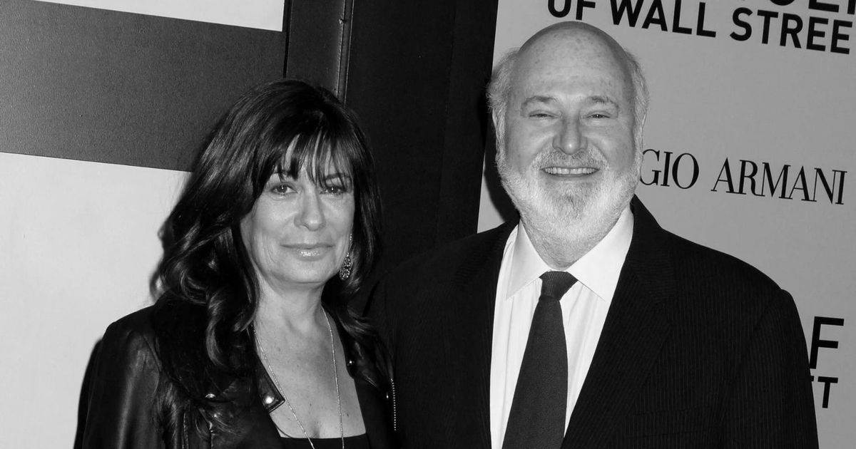 Dos personas en trajes elegantes posan en un evento formal con un fondo con texto impreso en blanco y negro.