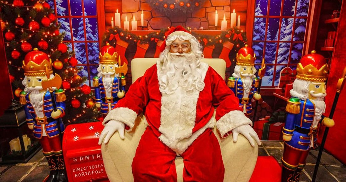 Santa Claus sentado en una silla con nutcrackers y adornos navideños, árbol de Navidad y nieve de fondo.