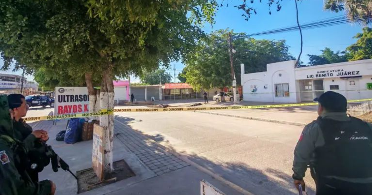 Escena urbana cautelosa en Villa Benito Juárez, Sinaloa, con árbol, cartel de servicios médicos y agentes de seguridad.