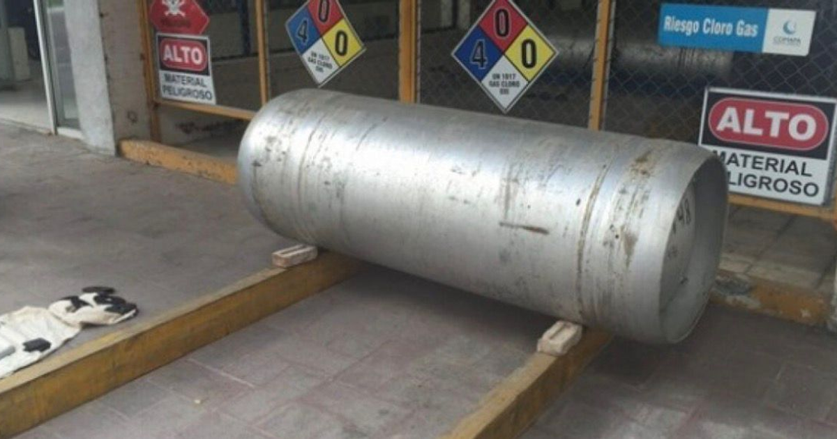 Cilindro de gas cloro con señalización de material peligroso, cerca de guantes y bolsa en zona química.