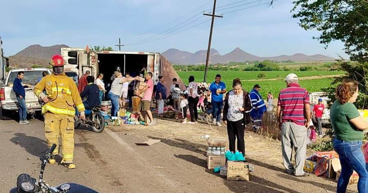 Personas y un bombero distribuyen suministros en un área rural con montañas al fondo, tras accidente de tráiler en Mocorito, Sinaloa.