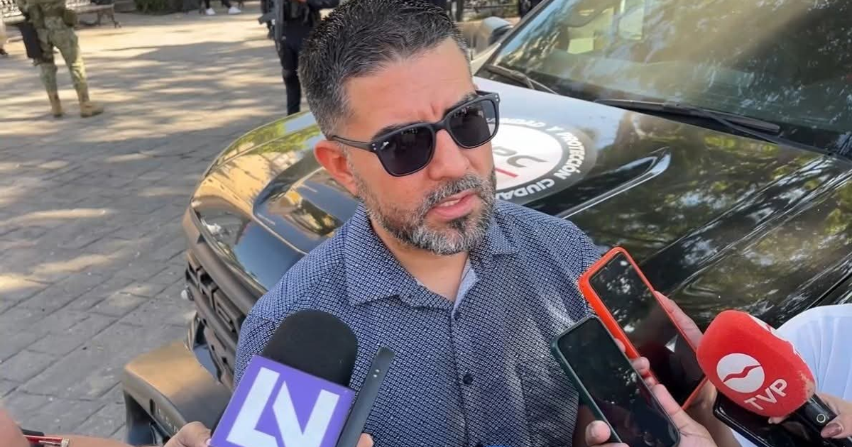 Hombre con gafas oscuras y barba, en rueda de prensa con periodistas, frente a una camioneta institucional