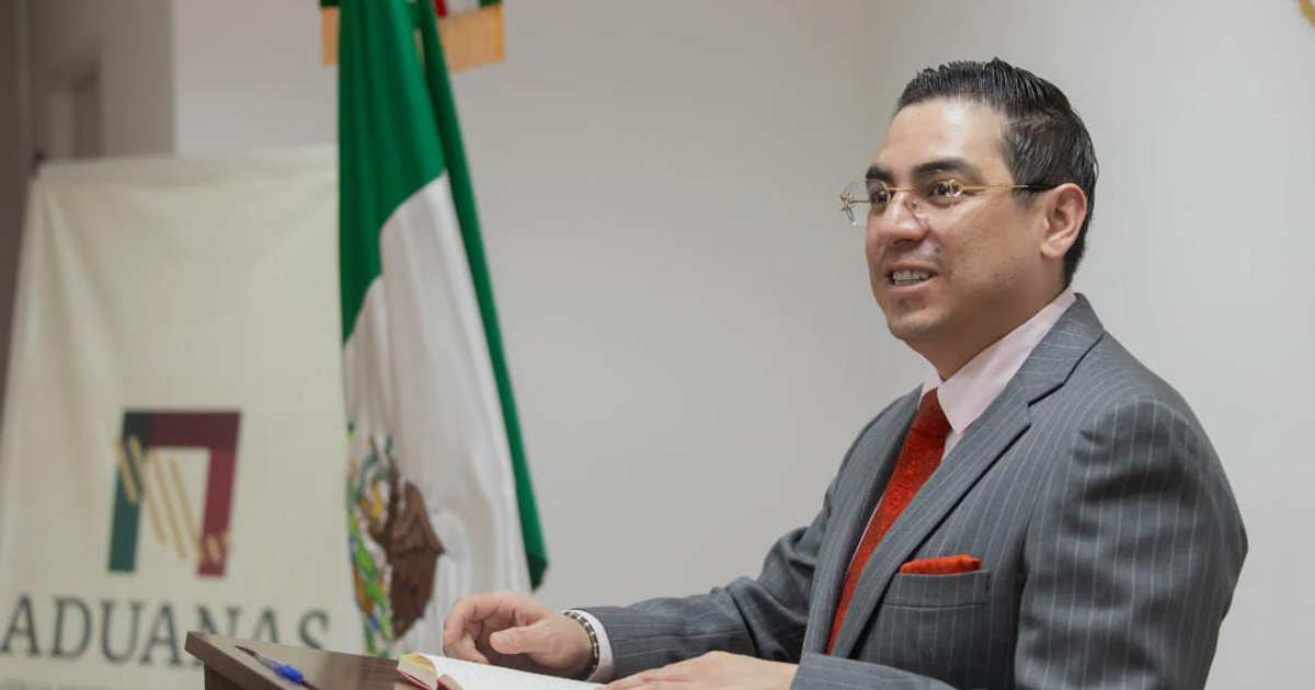 Alex Tonatiuh Márquez Hernández en conferencia con cartel de aduanas y bandera