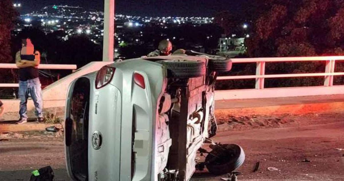 Automóvil Hyundai i10 volcado en un puente urbano de Culiacán durante la noche, con un individuo de pie cerca del vehículo y las luces de la ciudad de fondo.
