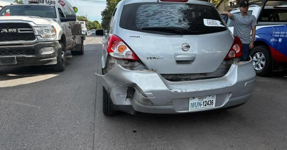 Nissan Versa dañado tras accidente en Mochis, Sinaloa, con camioneta blanca cercana y persona observando.