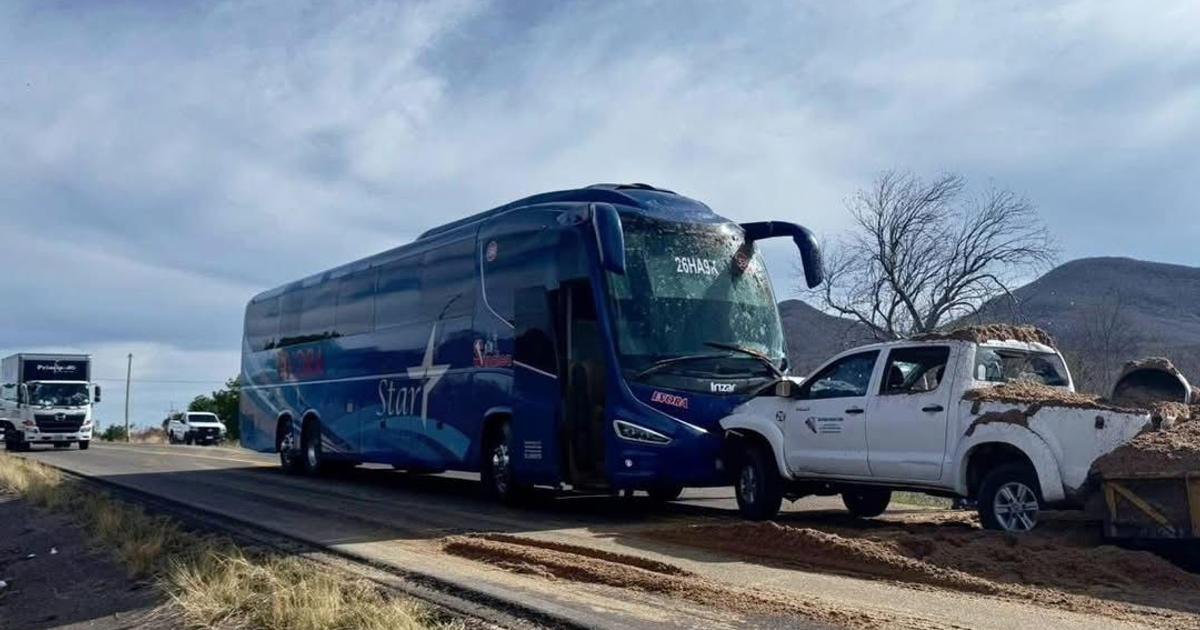 Autobús azul detenido en carretera, junto a una pickup blanca con carga de arena, cielo nublado y fondo montañoso.