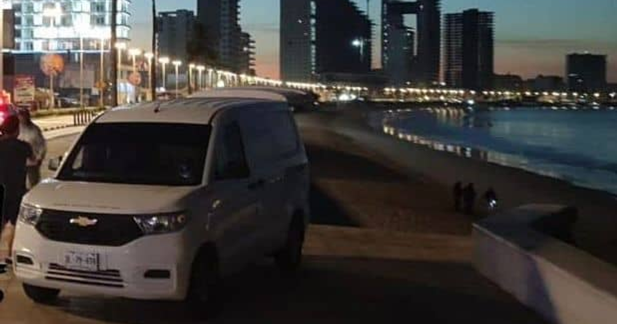 Van blanca estacionada en paseo de playa nocturna en Mazatlán, con luces y personas de fondo.