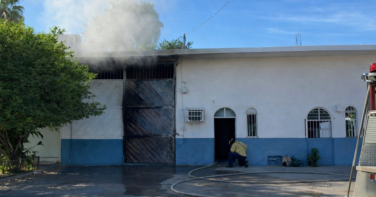 Edificio en llamas siendo controlado por bomberos en Los Mochis, Sinaloa, con humo saliendo de la parte superior y mangueras extendidas en el suelo.