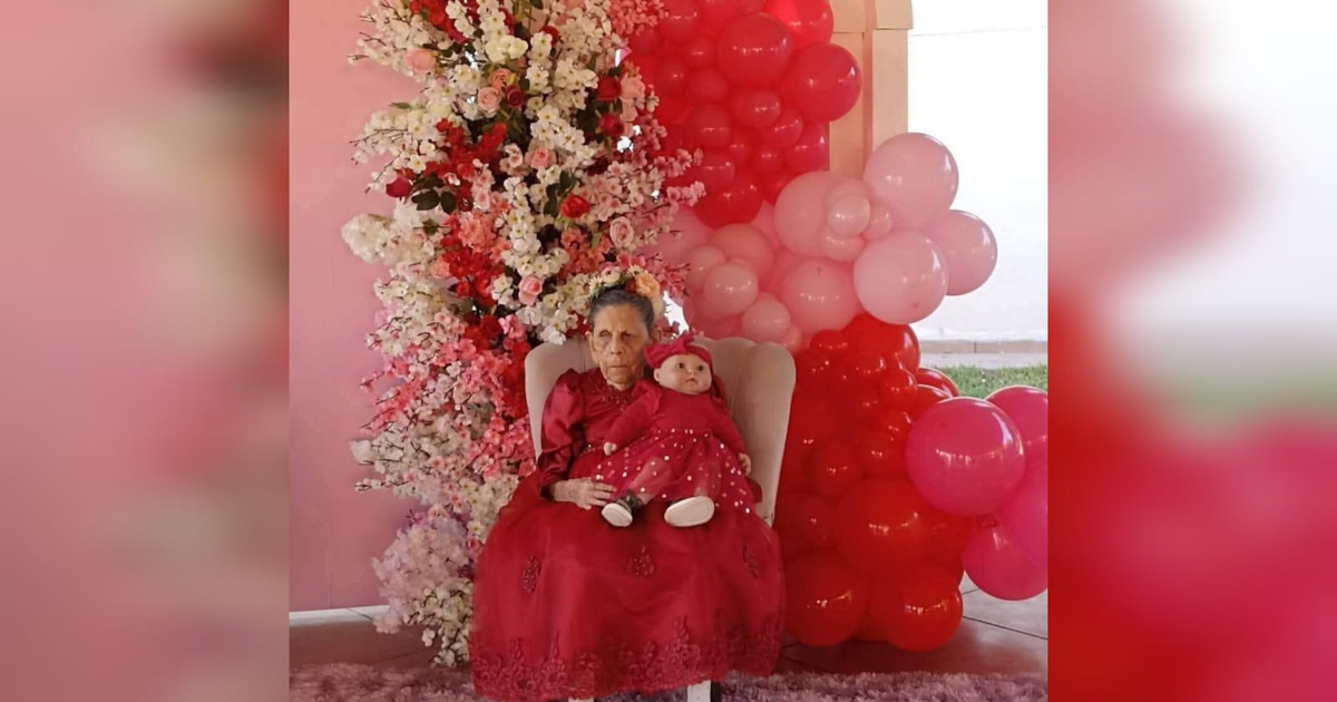 Una anciana y una muñeca en un entorno festivo de flores y globos rojos y rosas, homenaje a La Marillita de Guasave.