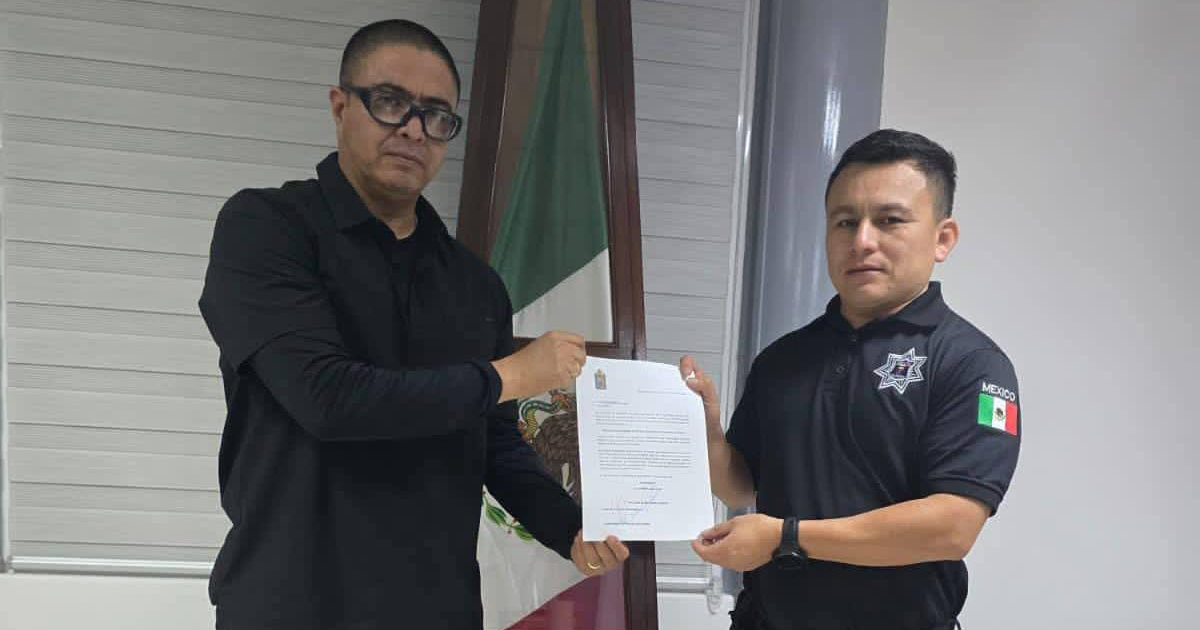 Dos hombres sosteniendo un documento en un acto oficial con bandera de México de fondo