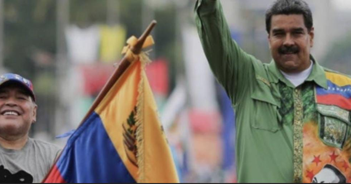 Dos hombres en un evento al aire libre, uno sosteniendo una bandera, otro saludando.