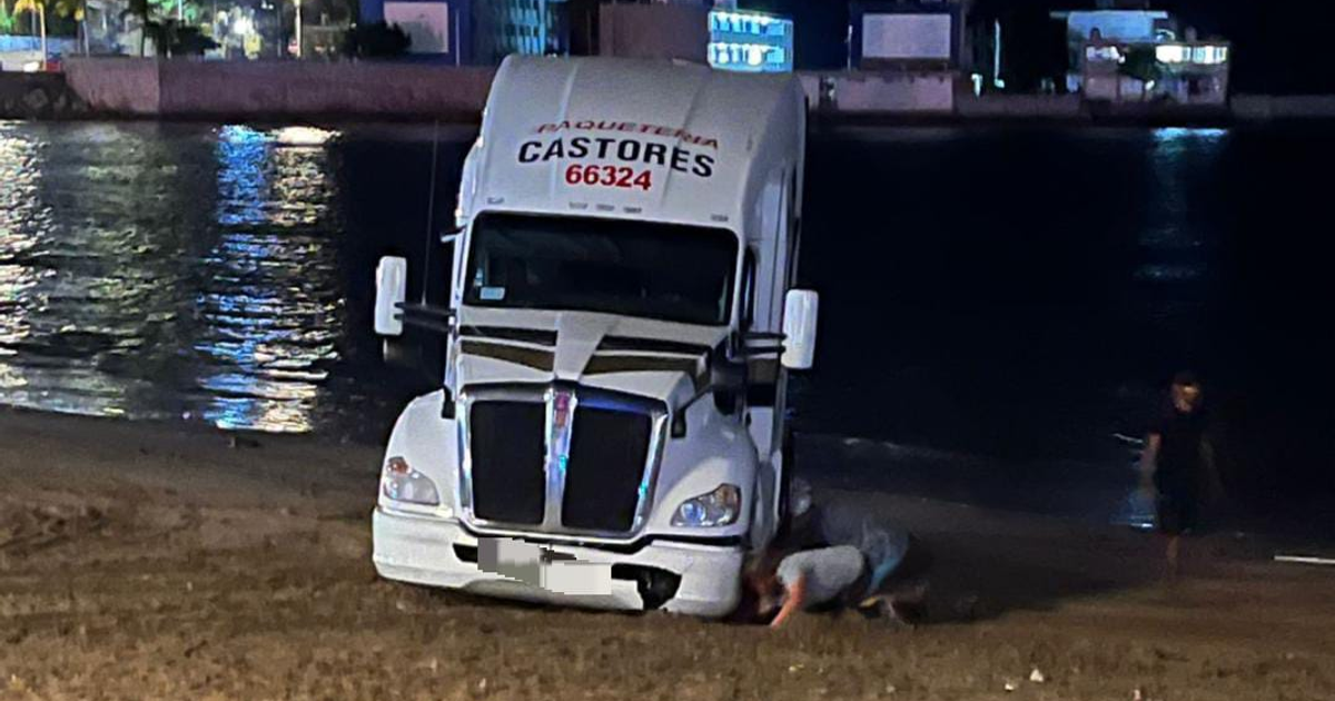 Tractocamión grande, varado en la playa de Mazatlán cerca de avenida Paseo Claussen, con personas y luces urbanas de fondo.