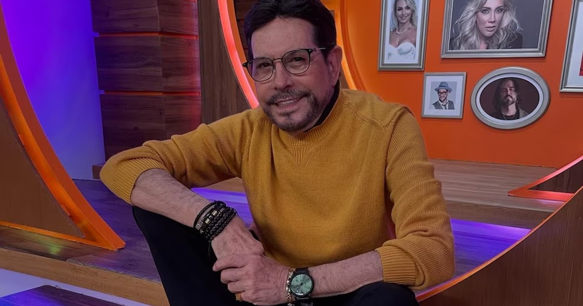 Juan José Origel sonriendo en set de TV con suéter amarillo y ambiante colorido.