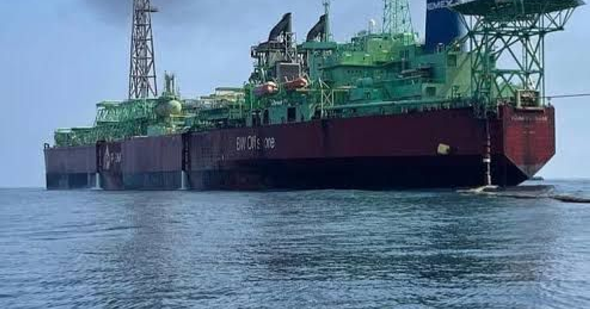 Buque de perforación petrolera anclado, estructuras verde y roja, plataforma en popa, cielo nublado.