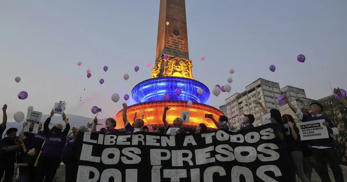 Manifestantes en Venezuela con pancartas y globos frente a un monumento iluminado, exigiendo libertad para presos políticos.