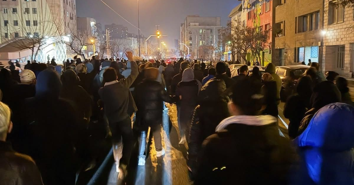 Grupo de personas vestidas con abrigos durante protestas nocturnas en Irán, con edificios y luces urbanos de fondo.