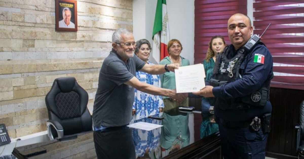 Un hombre de cabello gris entrega documentos a un oficial de policía en una oficina con una bandera mexicana y un retrato en la pared.