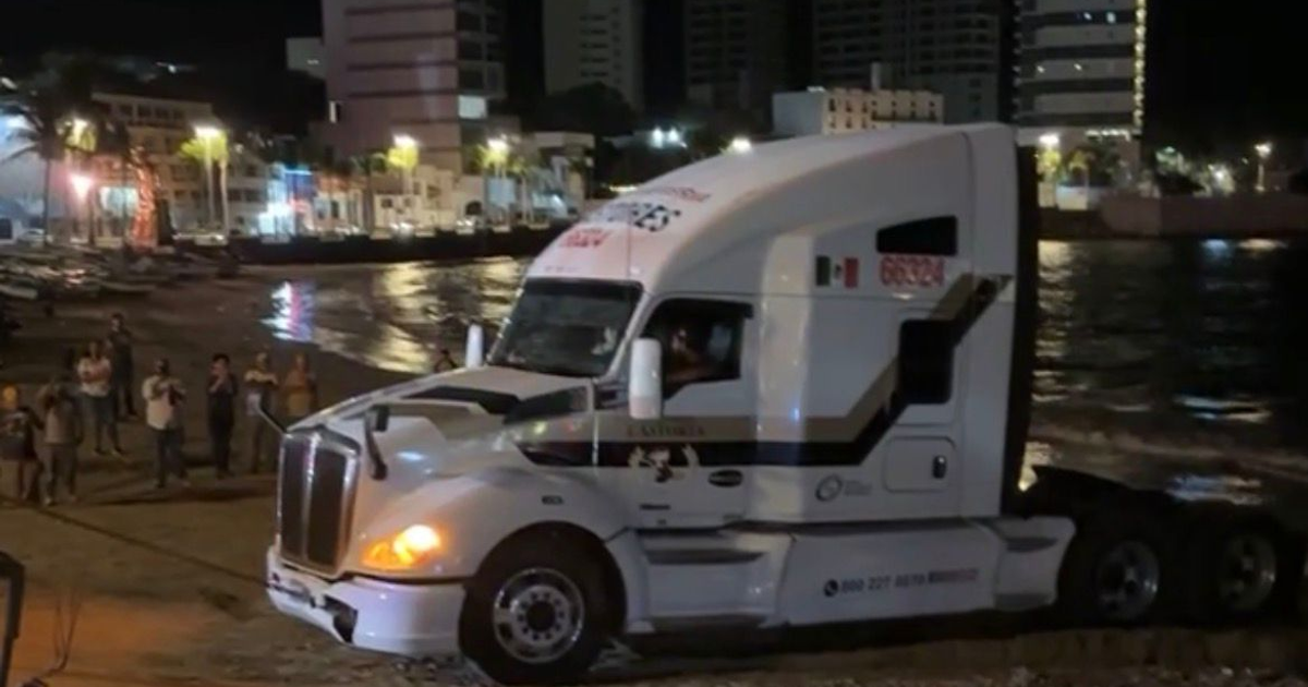 Camión de carga varado en Playa Norte, Mazatlán, con palmeras y luces nocturnas de fondo.